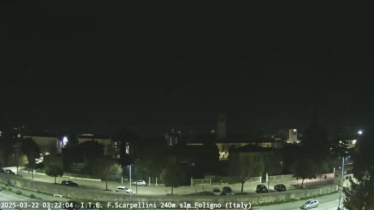 A nighttime view of a town with sparsely parked cars along a roadway, under a clear, dark sky.