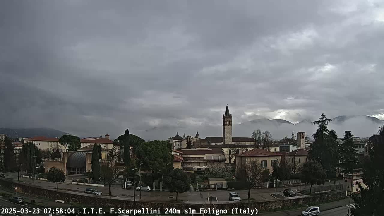 A cloudy, overcast day reveals a town with buildings, trees, and a tall bell tower, with low-lying fog or clouds partially obscuring distant mountains.