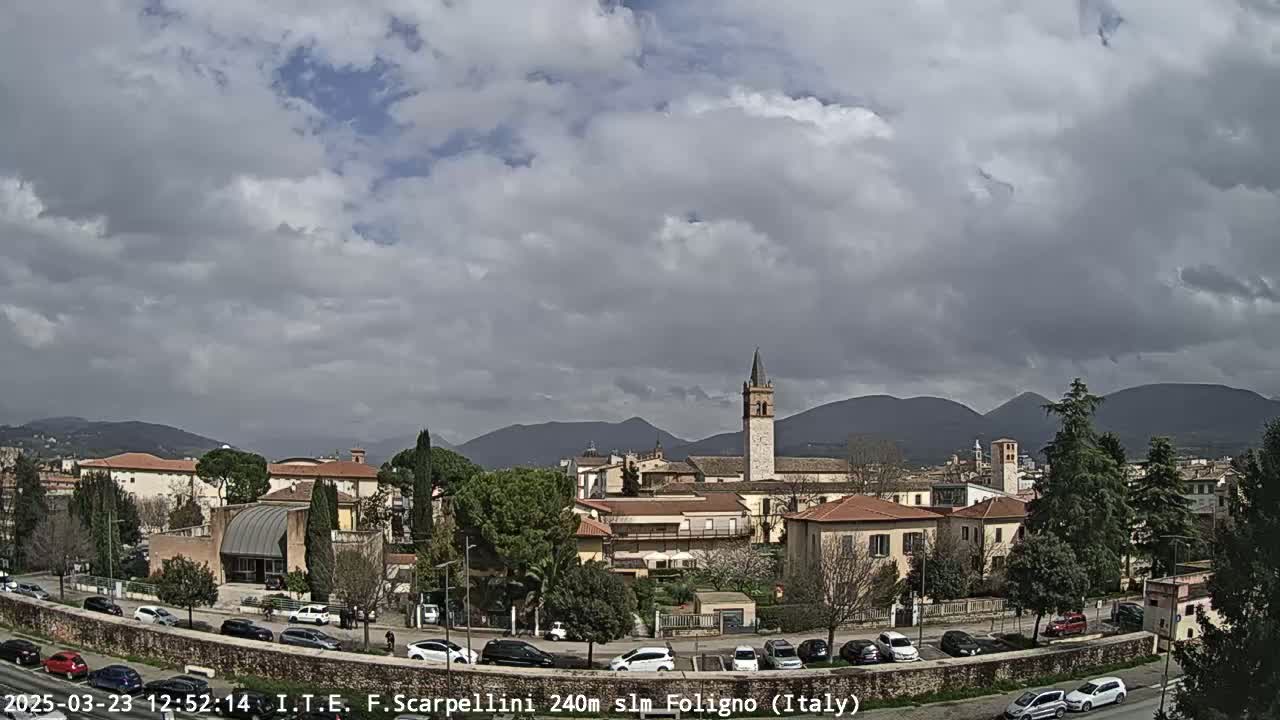 A partly cloudy day reveals a cityscape of low buildings, a tall church tower, and distant mountains.