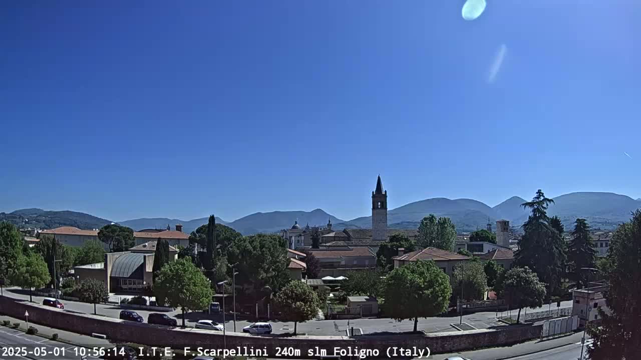 A sunny day reveals a picturesque Italian town nestled amongst rolling hills and mountains, with a prominent bell tower standing tall in the center.