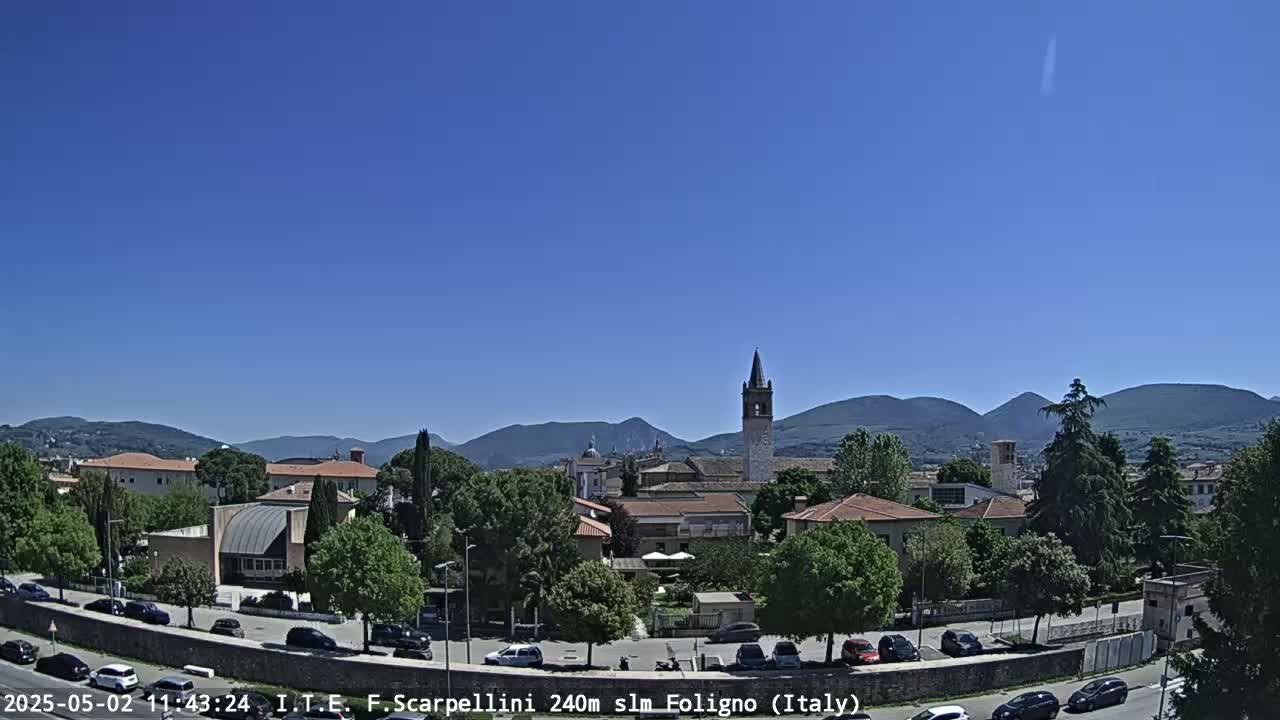 A sunny day reveals a town nestled among green hills and mountains, with cars parked along a road in the foreground.