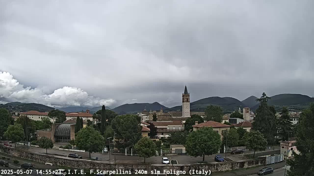 A cloudy day reveals a town nestled among hills, featuring buildings with red-tiled roofs and a tall, slender tower.