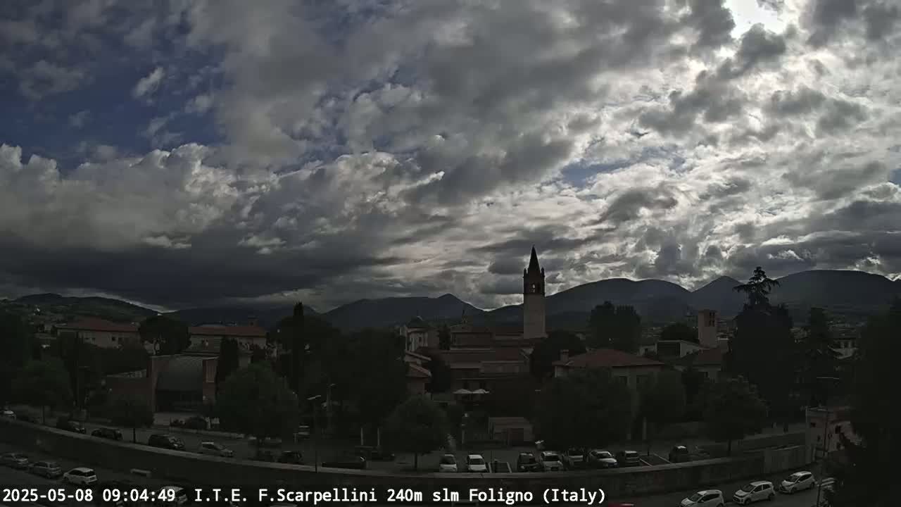 A town nestled in a valley between mountains is partially obscured by a mostly cloudy sky.
