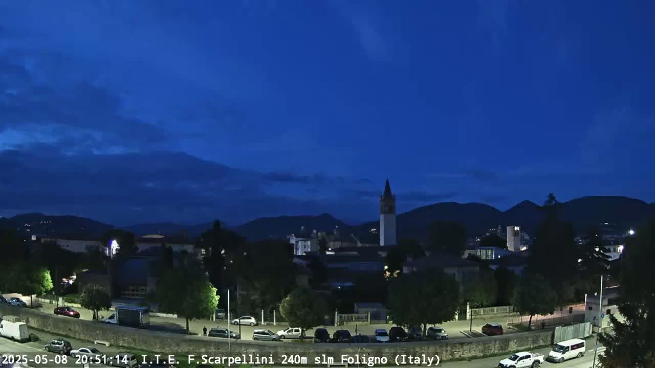 A nighttime view of a town nestled in a valley, under a mostly clear, dark blue twilight sky, with mountains in the background.