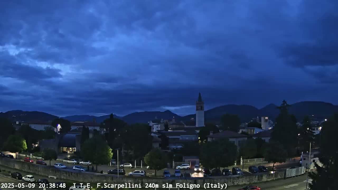 Under a dark blue, cloudy sky, a small town nestled at the base of mountains is illuminated at night.