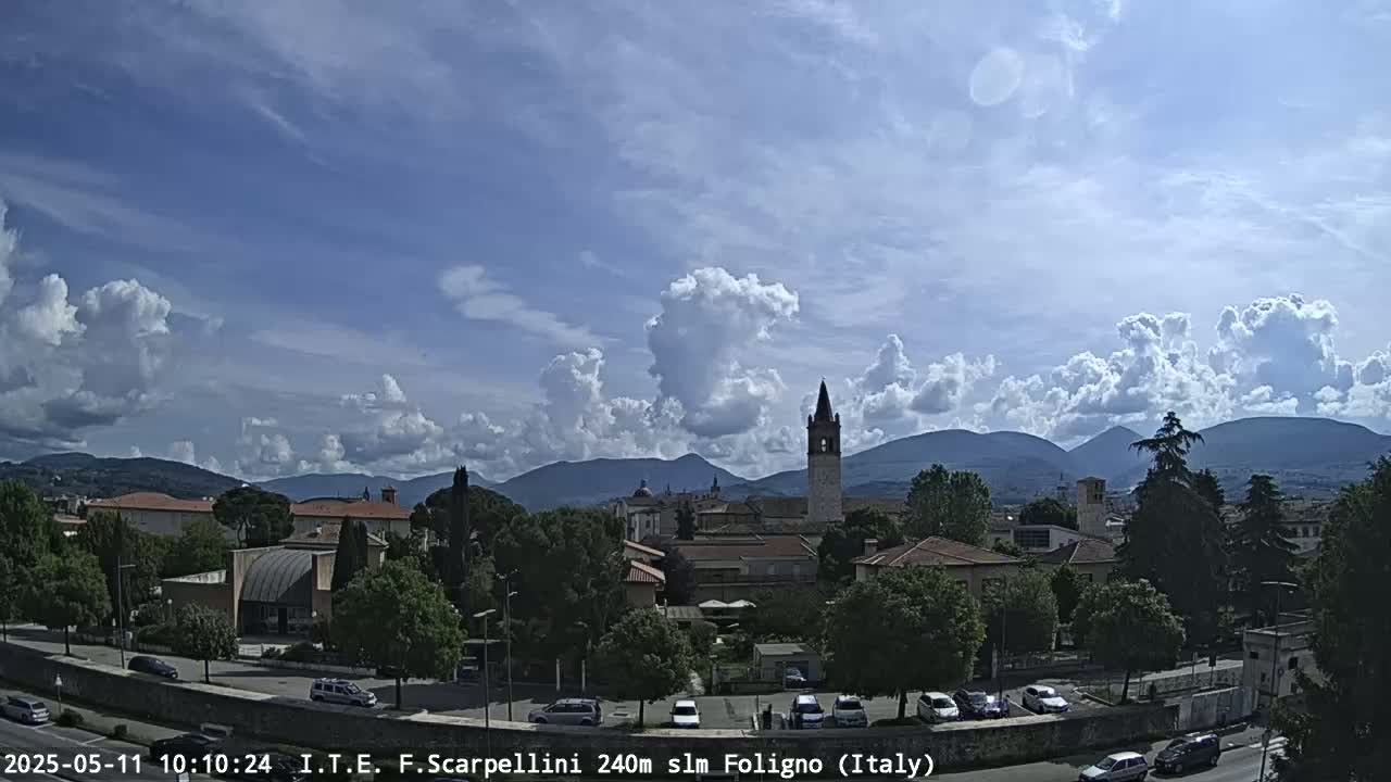 A partly cloudy day reveals a town nestled in a valley, with mountains in the distance and a tall bell tower prominent in the cityscape.