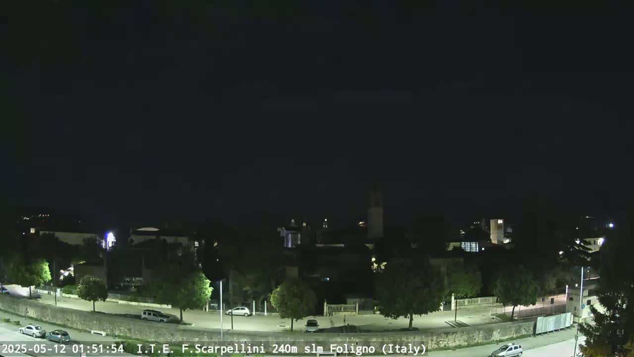 A nighttime, long shot of a town with a few cars parked and driving along a curved road under a dark sky.