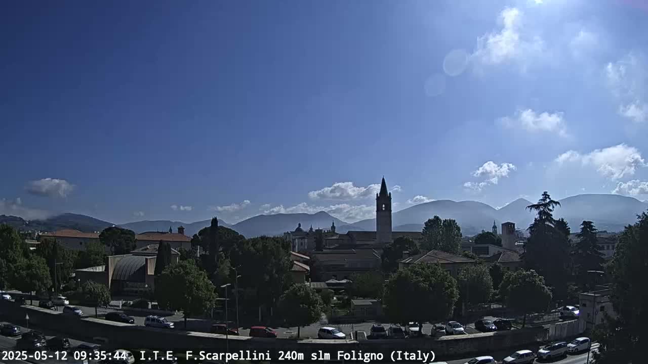 A sunny day reveals a town nestled among low mountains, with a tall church steeple prominent in the center, and cars parked along a road in the foreground.