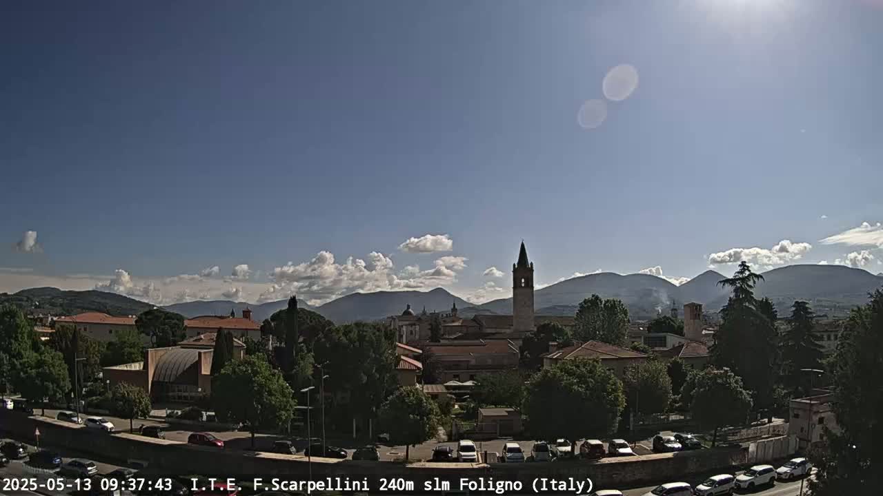 A sunny day reveals a picturesque Italian town nestled among hills, with a tall bell tower prominent in the cityscape.