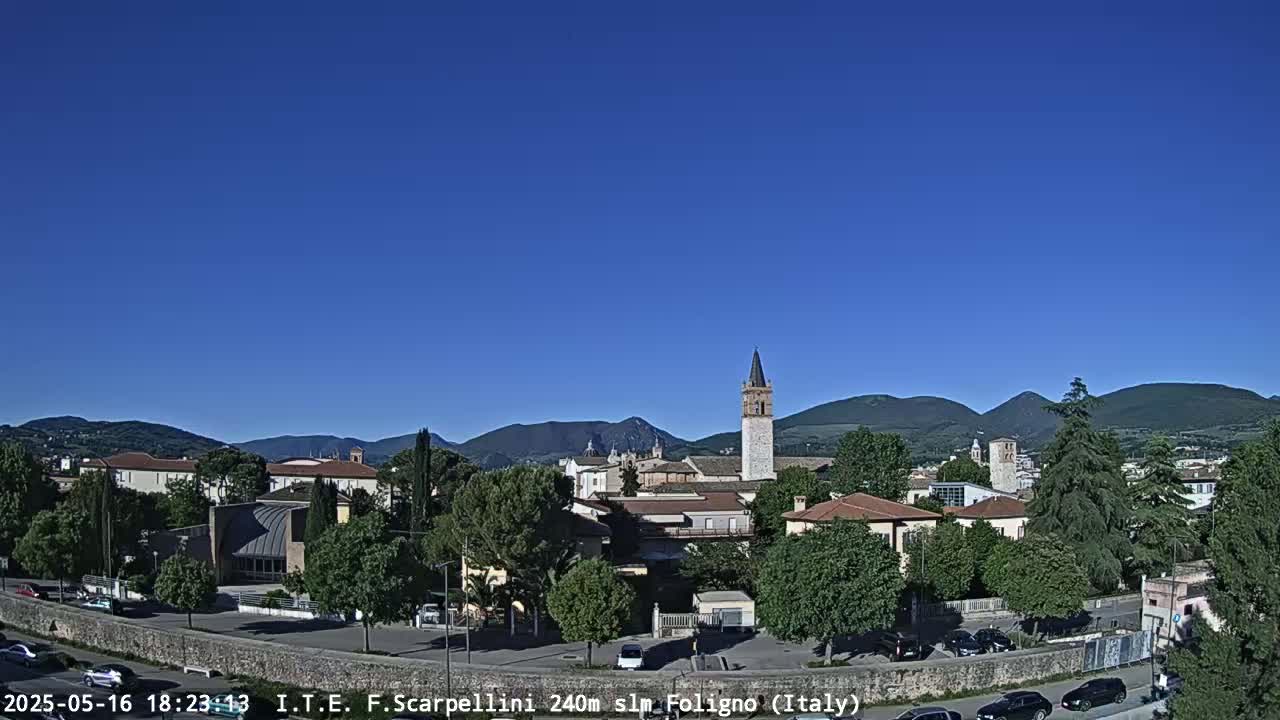 A town nestled in a valley under a clear blue sky, with mountains in the distance and cars parked along a wall.