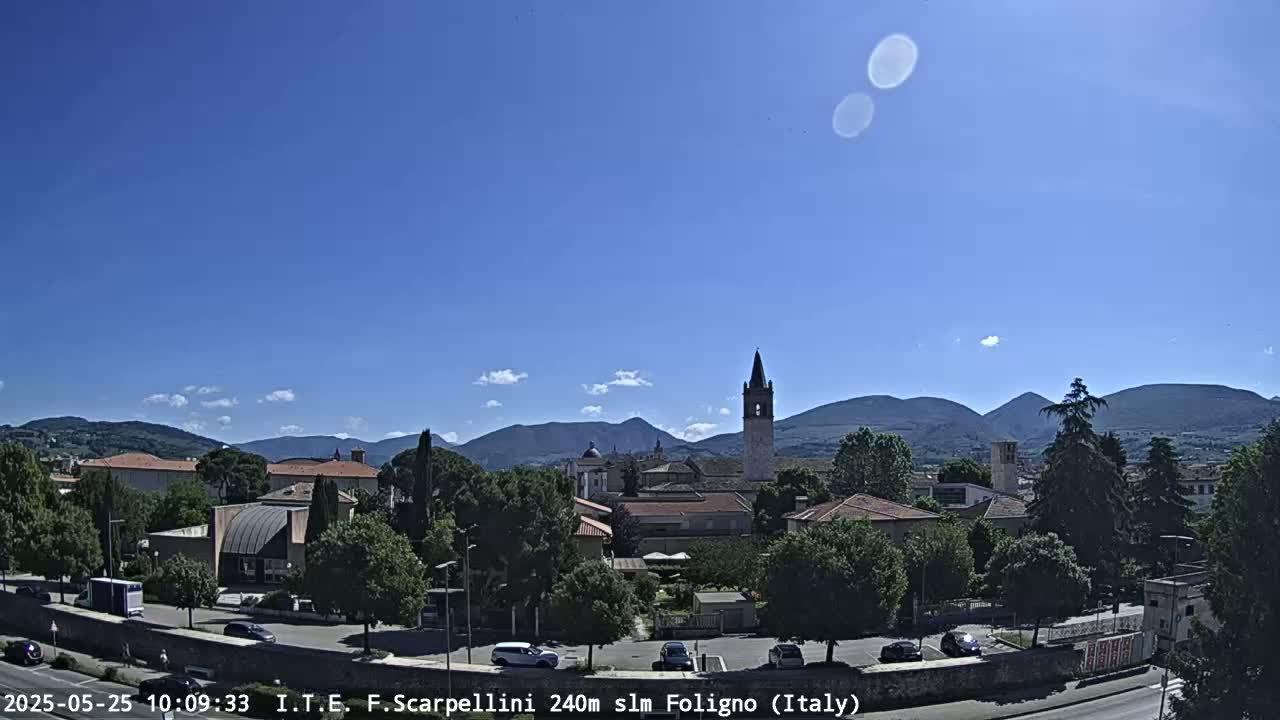 A sunny day reveals a picturesque Italian town nestled among rolling hills and mountains, with a prominent bell tower standing tall amongst the buildings.