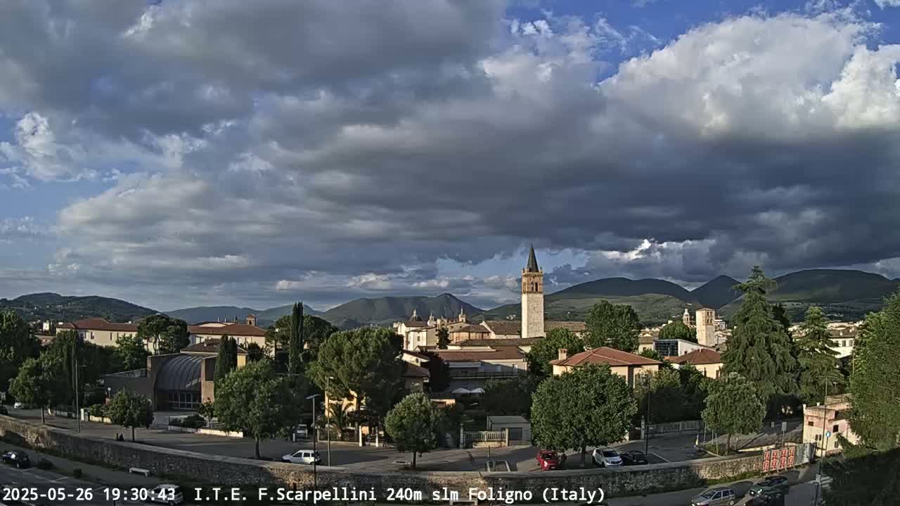 A partly cloudy sky hangs over a town nestled in a valley between mountains.