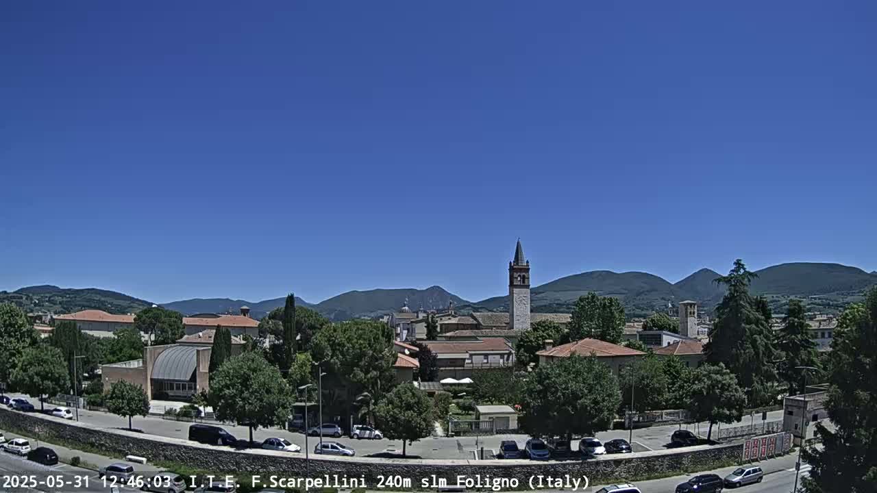 A clear, sunny day reveals a town nestled in a valley, surrounded by mountains, with a tall bell tower prominently featured.
