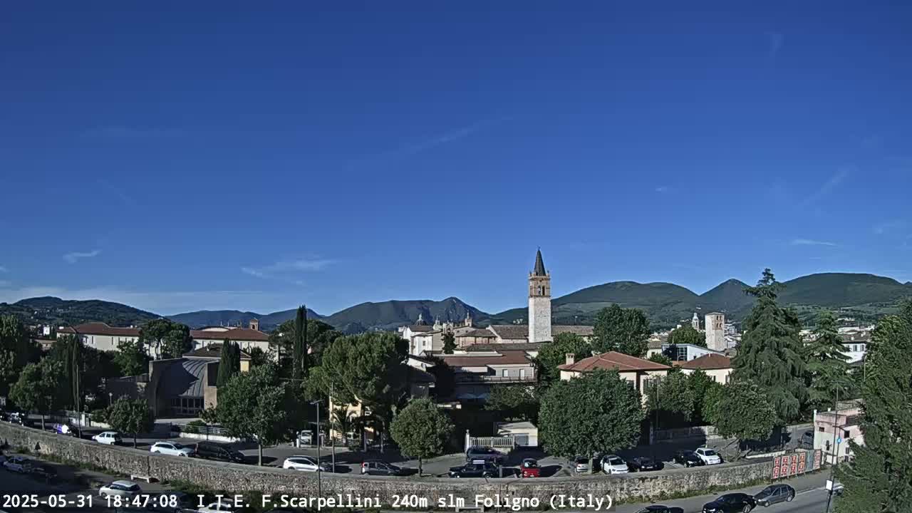 A picturesque Italian town, nestled among rolling hills under a clear blue sky, features a prominent bell tower and several buildings.