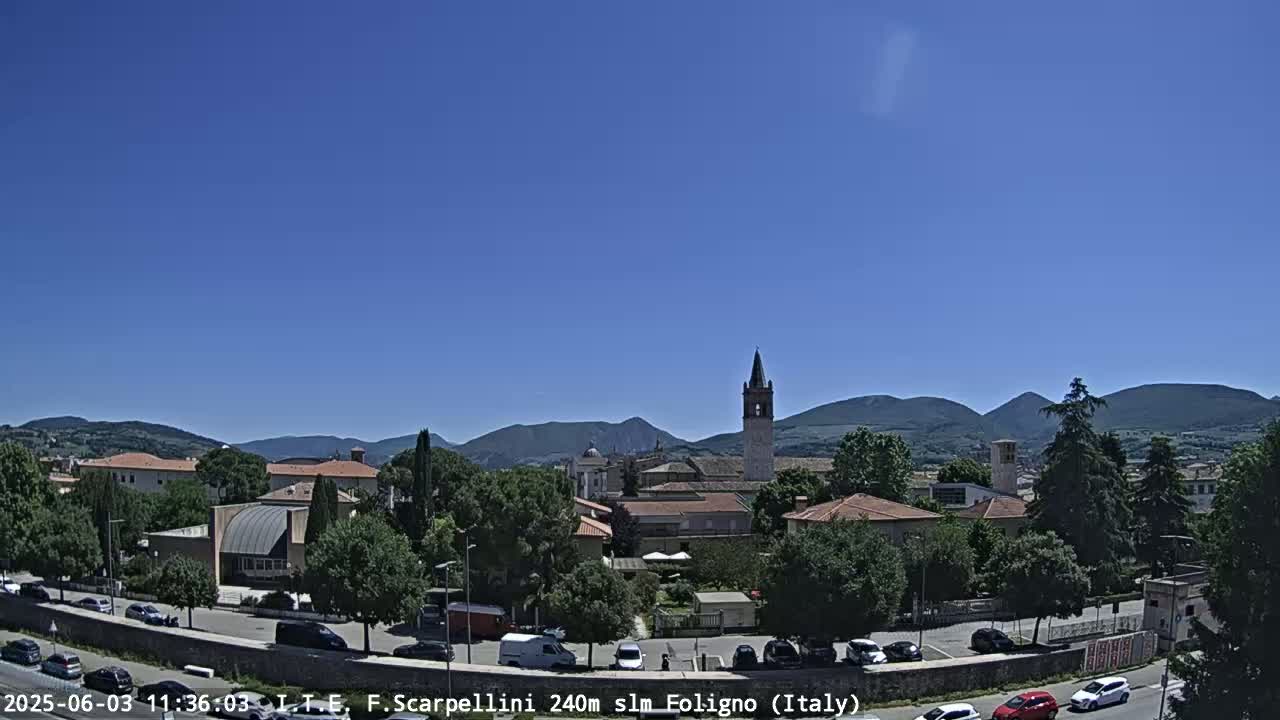 A sunny day reveals a town nestled amongst green hills and mountains, with a tall bell tower prominently featured in the cityscape.