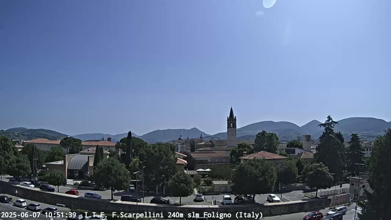 A sunny day reveals a town nestled among green hills and mountains, with a tall tower standing prominently in the center.