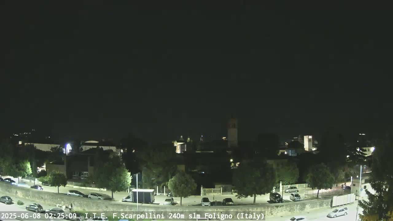 A nighttime, clear-sky view of a town with cars parked along a road, and buildings and trees visible under artificial light.