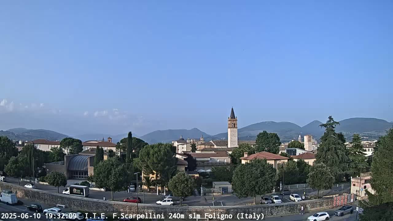 A clear, sunny day reveals a town nestled in a valley, with mountains in the background and cars parked along a road in the foreground.