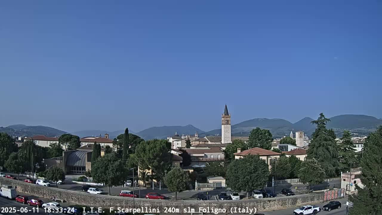 A clear, sunny day reveals a picturesque Italian town nestled among rolling hills, with a tall, slender tower as a prominent feature, and cars parked along a wall in the foreground.