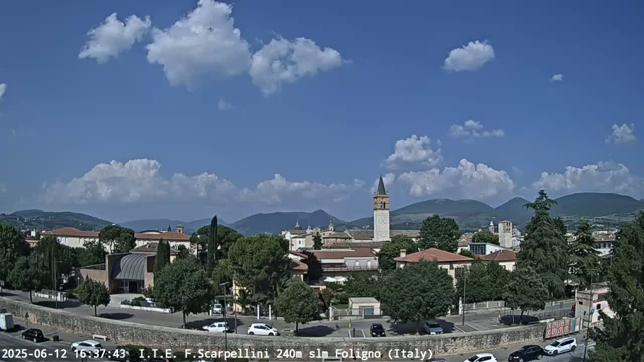 A partly cloudy blue sky overlooks a town with low buildings, trees, and a tall tower, set against a backdrop of distant mountains.