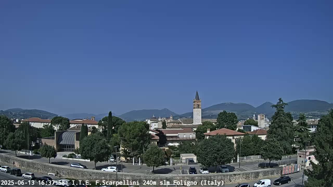 A sunny day reveals a town nestled among mountains, with a tall, slender tower standing prominently among low buildings and green trees.