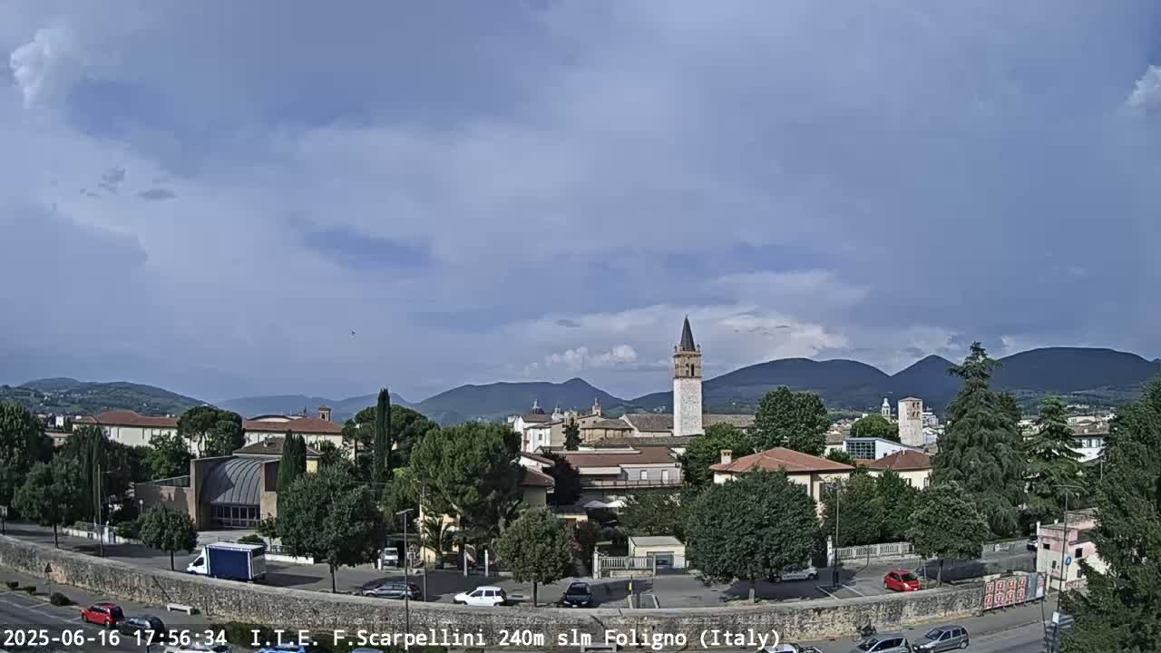 A partly cloudy day reveals a town nestled amongst green trees and hills, with a tall tower prominently featured in the center.