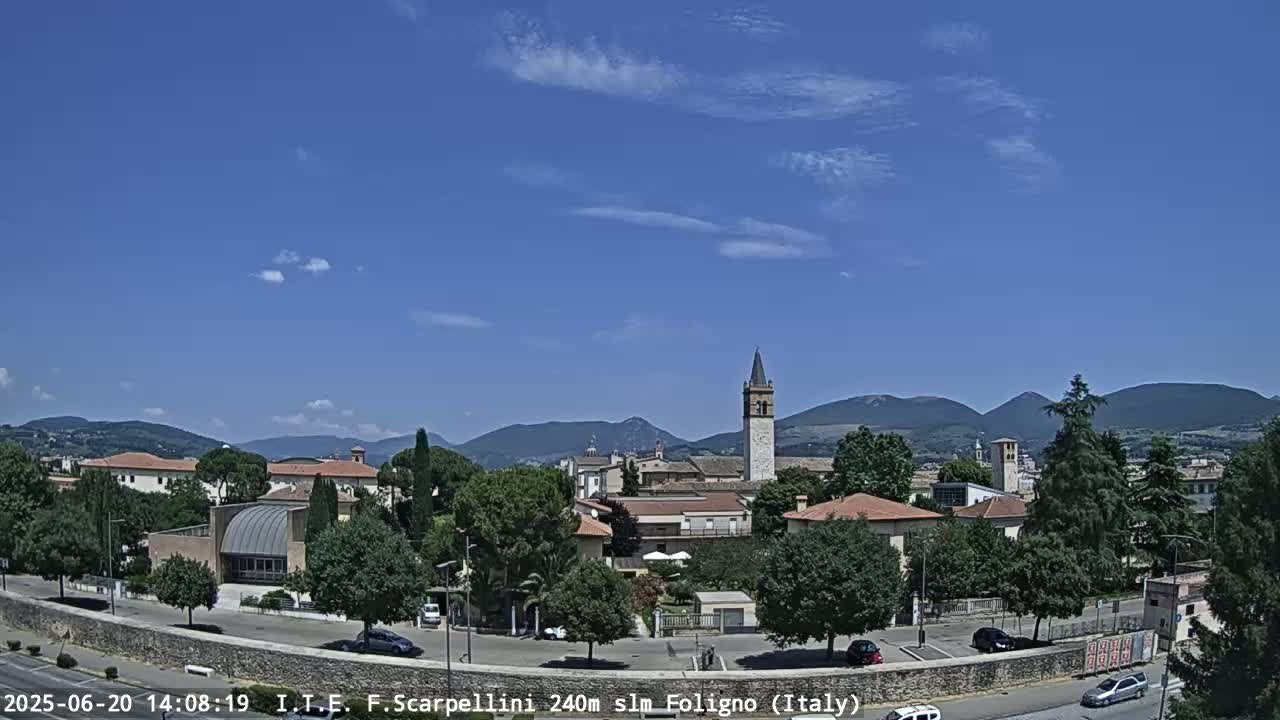 A sunny day reveals a town nestled among hills, featuring buildings with terracotta roofs and a prominent bell tower, with a few cars parked on a street below.
