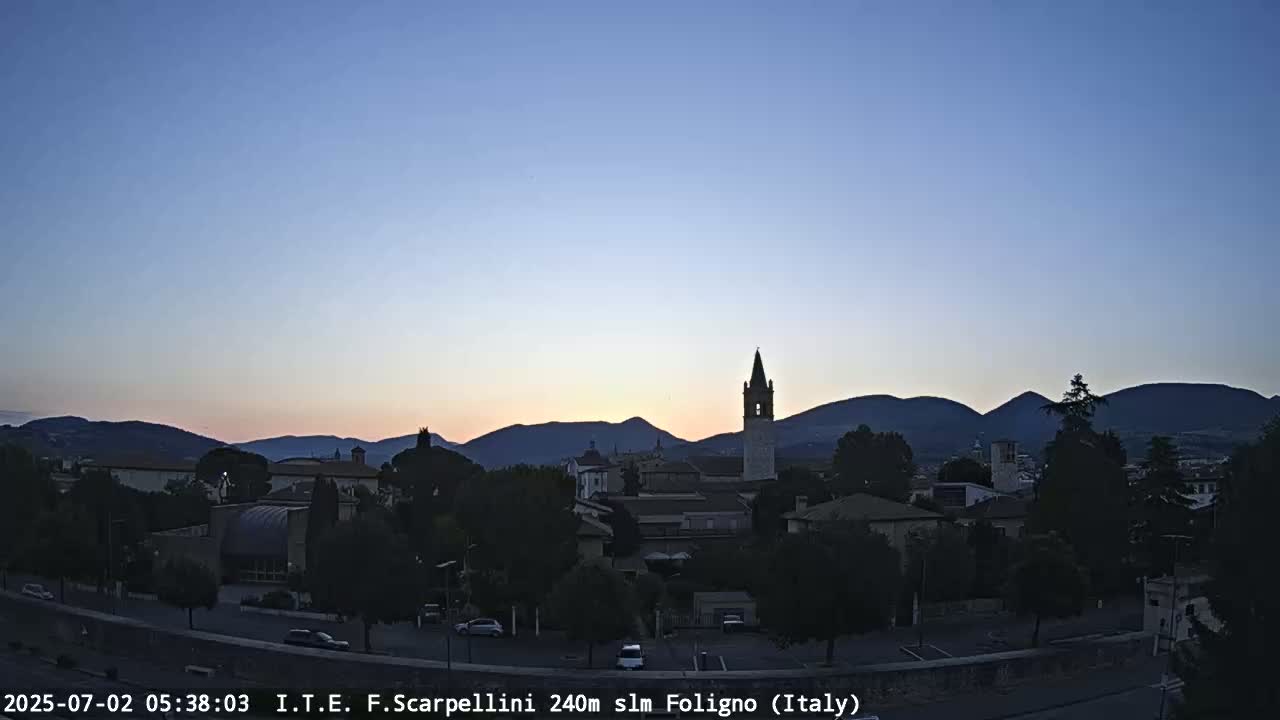 A pre-dawn view of a town nestled in a valley, silhouetted against a clear, pale sky and mountains in the distance.