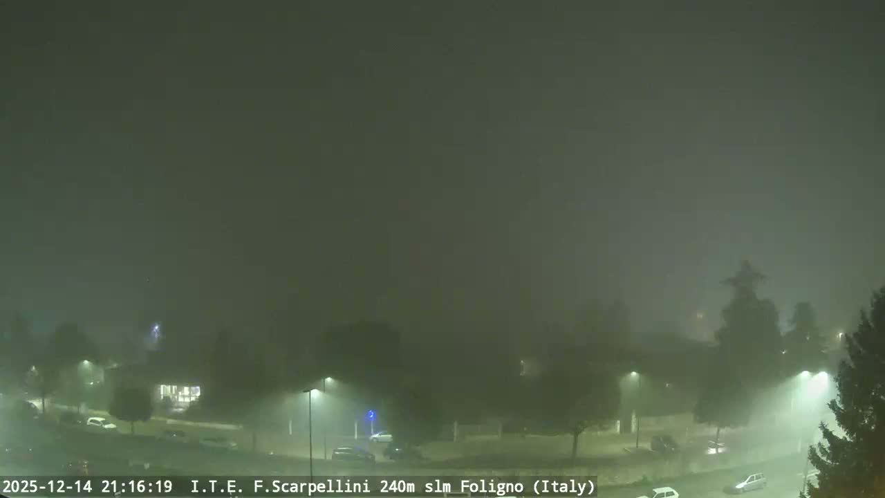 An expansive view of a European town on a uniformly gray, overcast day, featuring a mix of residential buildings, numerous trees, and streets lined with parked cars, all beneath a prominent church spire in the background.