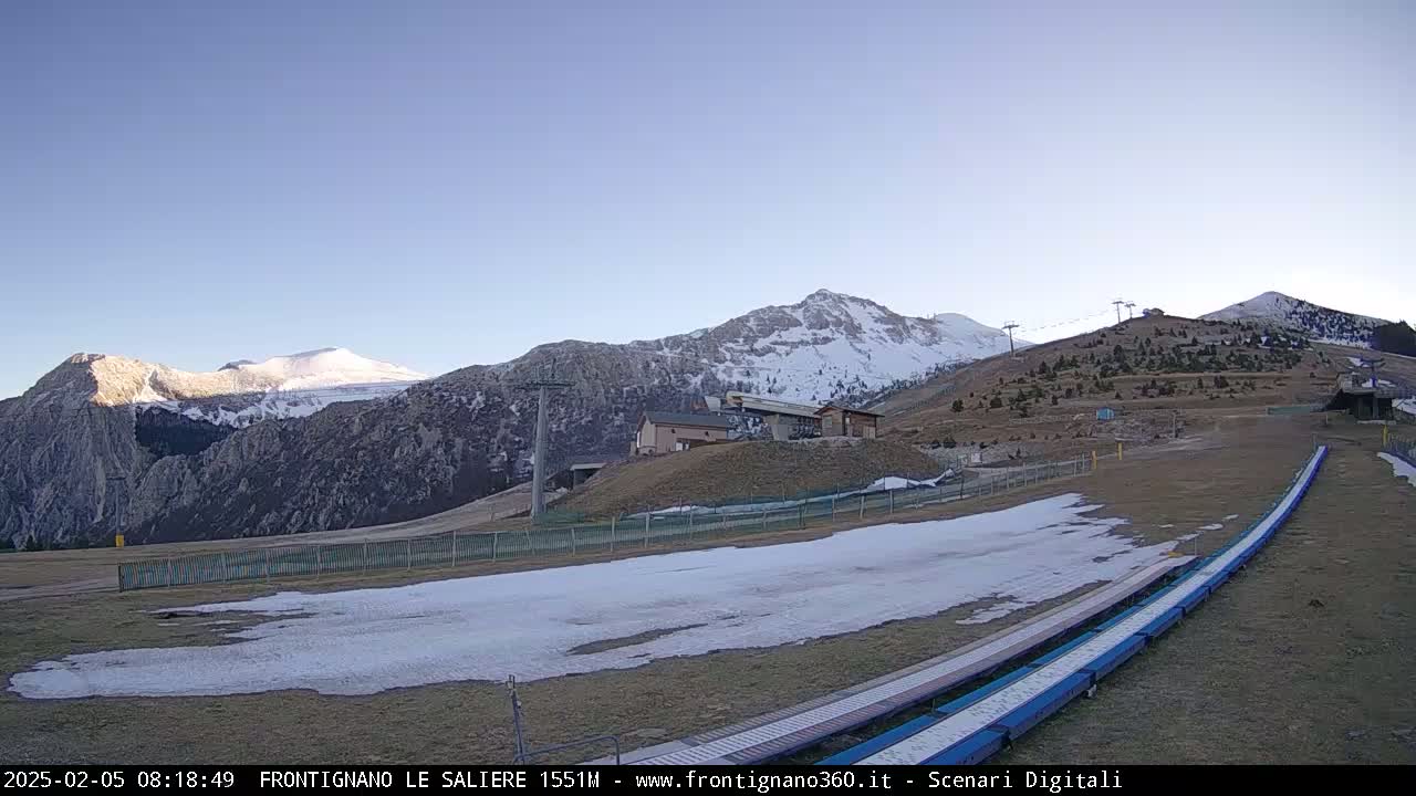 A ski slope with patches of snow sits below snow-capped mountains under a clear blue sky.
