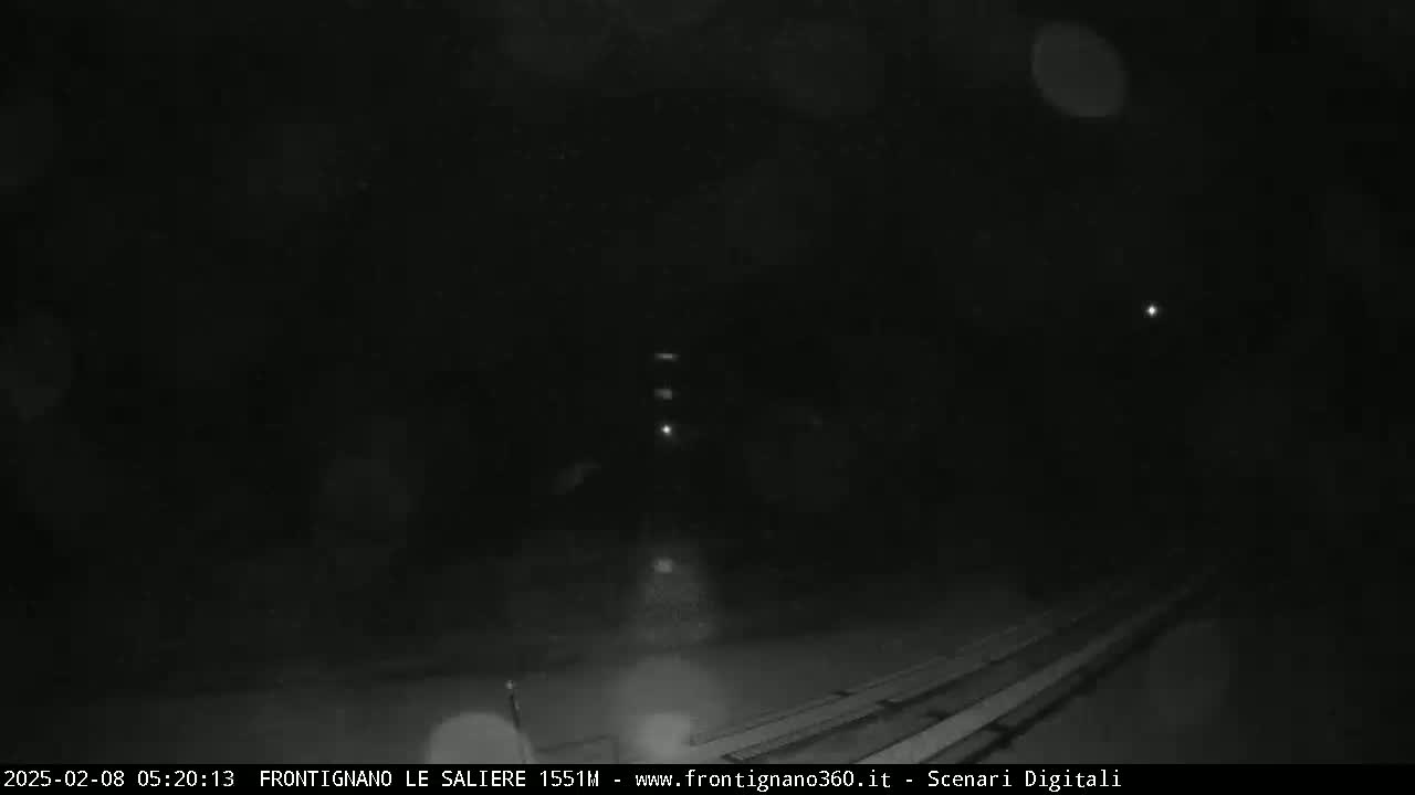 A nighttime, dark scene shows a few distant lights and a mostly obscured structure in the lower right corner, possibly under snowy or dusty conditions.