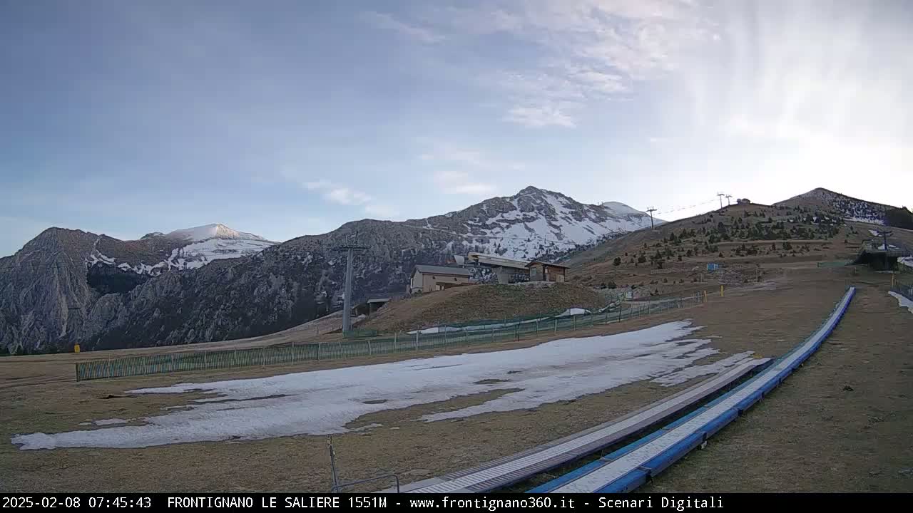 A mostly clear sky overlooks a snow-dusted mountain range with ski lifts and runs, showing patches of snow on the ground.