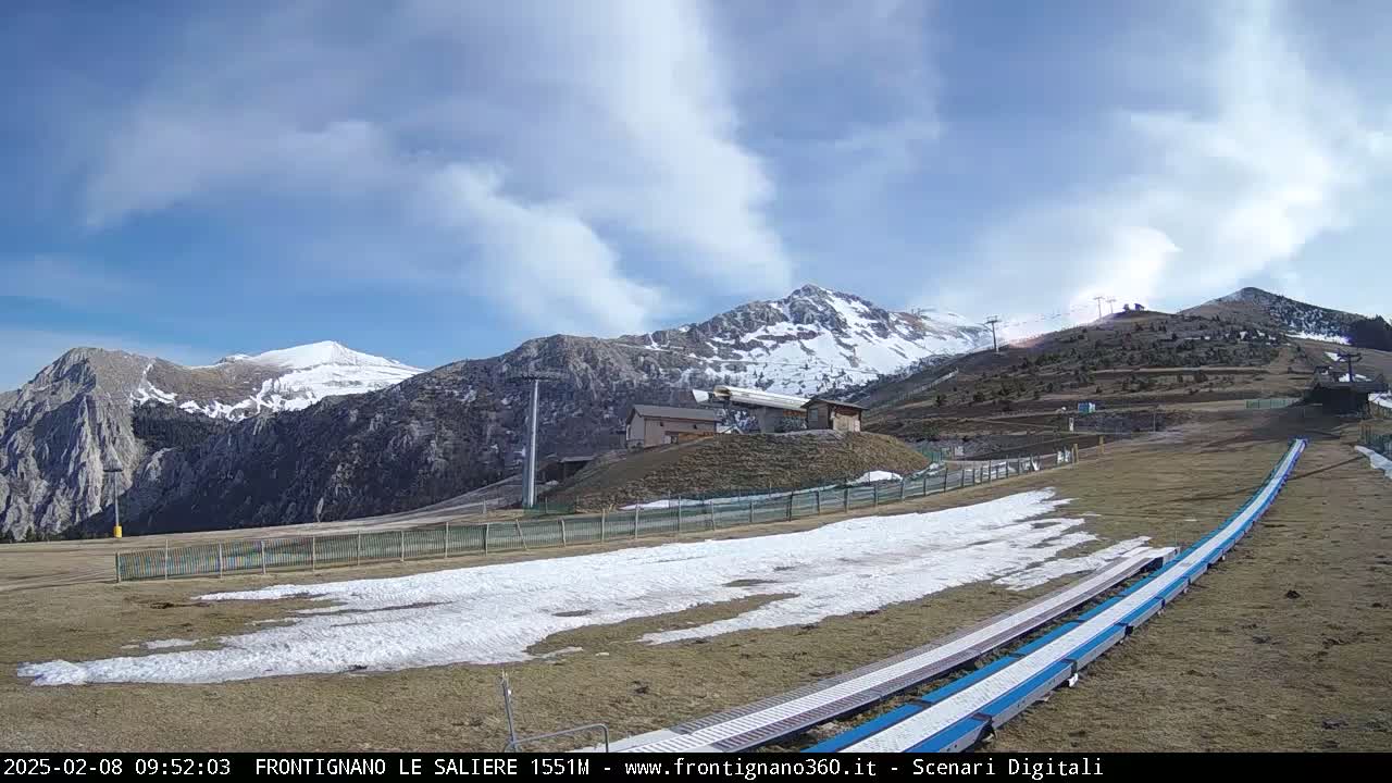 A mostly clear, sunny day reveals snow-dusted mountains and a ski slope with patches of snow and a snow-making machine.