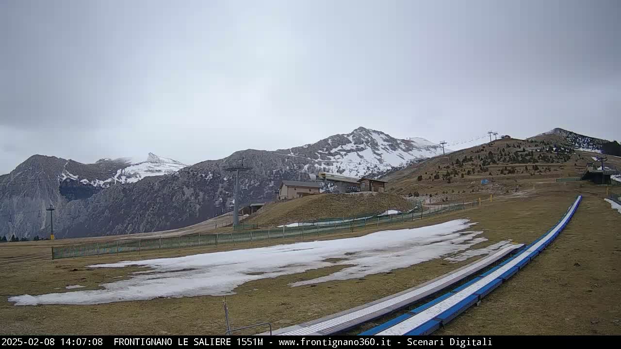 A snow-dusted mountain range is visible under an overcast sky, with a grassy area in the foreground containing patches of snow and a long, blue toboggan run.