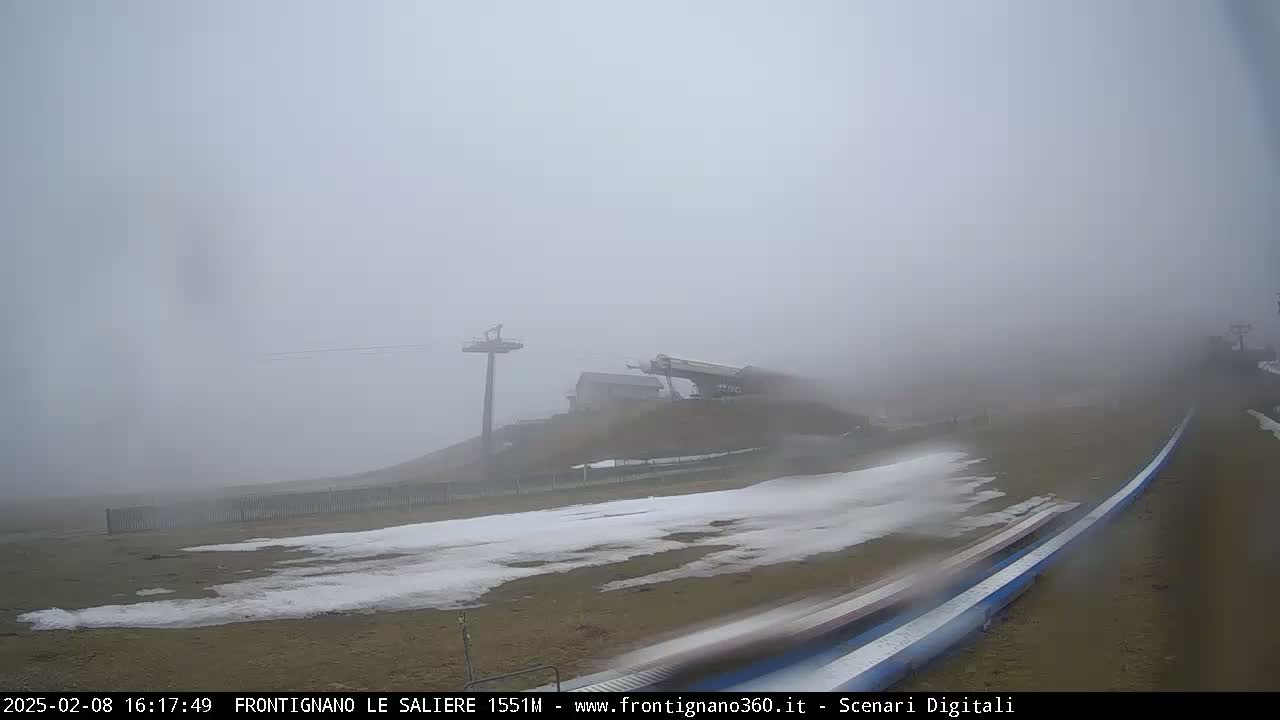 A foggy scene shows a ski lift and a snow-covered slope with a long, low structure extending from the right side of the image.