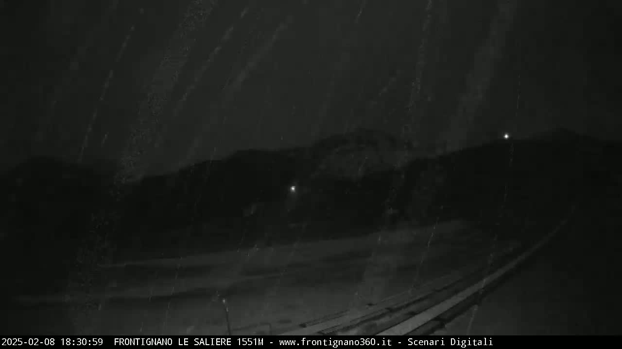 A nighttime, snowy scene shows a dark roadway leading toward dimly lit hills under a dark sky.