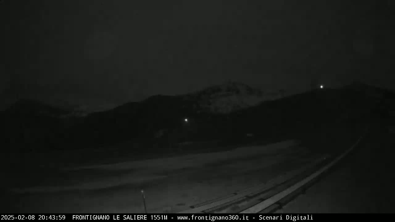 A nighttime, low-light image shows a dark, snow-covered mountain range in the distance, and a flat, dark expanse of land in the foreground with what appears to be a long, narrow structure at the bottom.