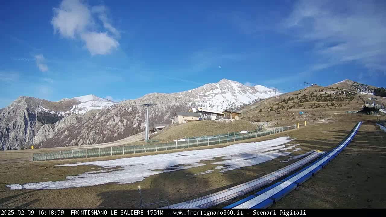 A partly sunny day reveals snow-capped mountains in the background, with a grassy foreground featuring a long, low snow tubing run.
