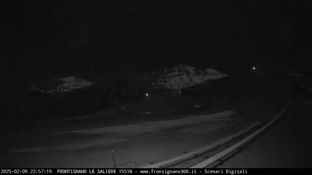 A nighttime, low-light view of a snow-covered landscape with bleachers and dimly lit areas in the distance.