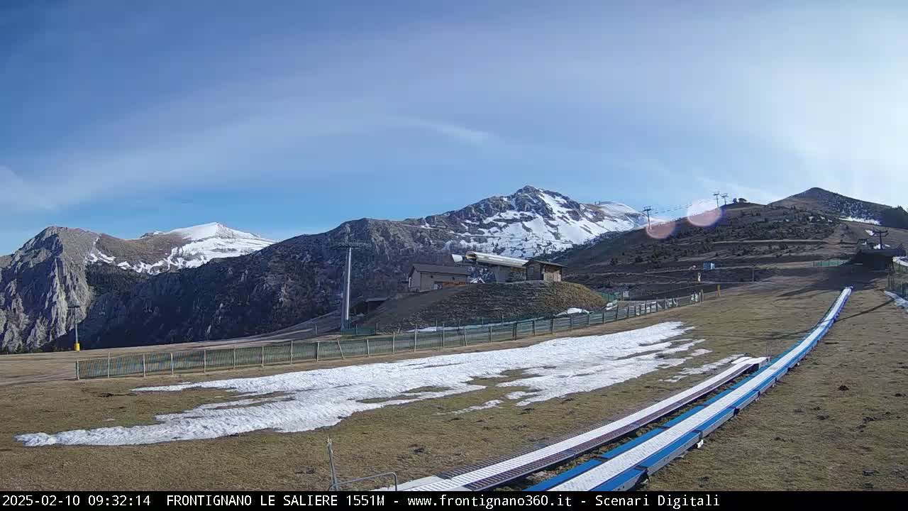 A sunny day reveals snow-capped mountains and a ski slope with a snowmaking machine, some patches of snow remaining on the ground.