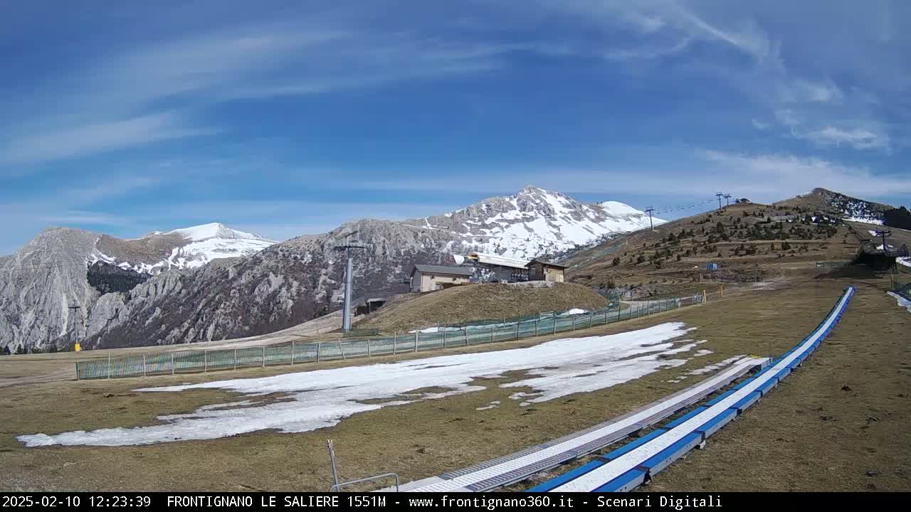A sunny day reveals a ski slope with patches of snow, a chairlift, and snow-capped mountains in the background.