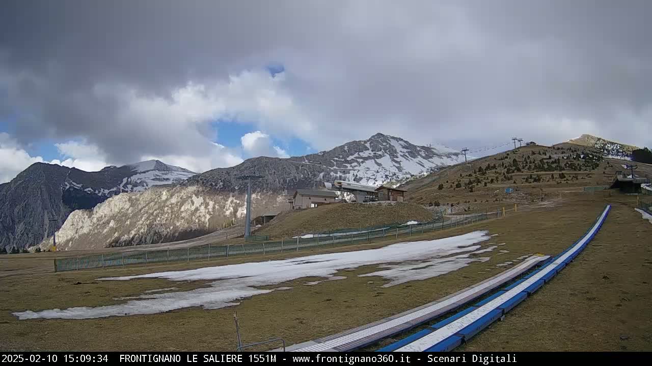 A partly cloudy day reveals snow-capped mountains and a ski lift area with patches of snow and brown grass.