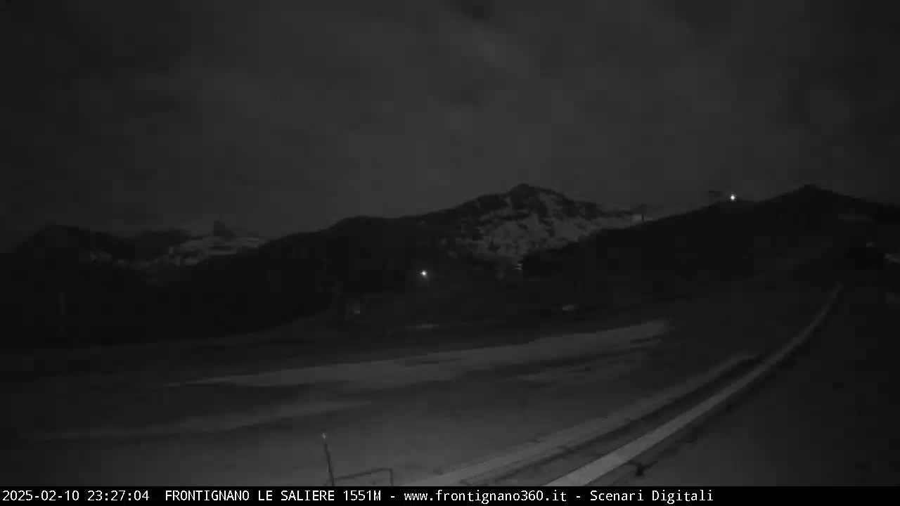 A nighttime, low-light view shows a dark road curving toward snow-dusted mountains under a cloudy sky.