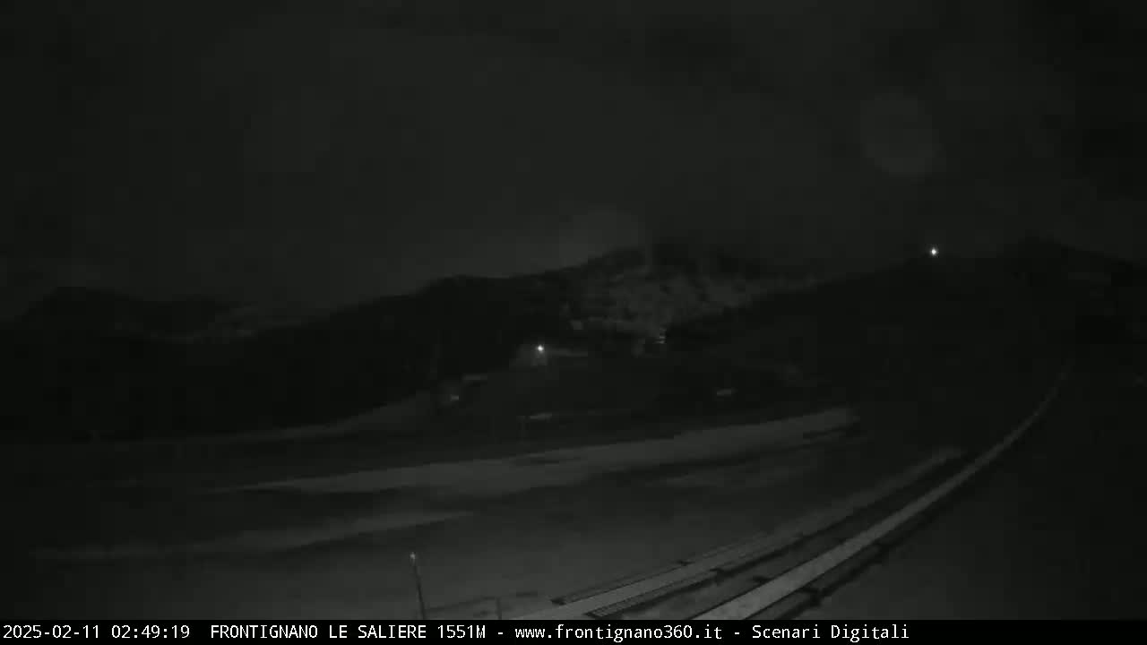 A nighttime, low-light view of a snow-covered landscape with hills in the background under a cloudy sky.