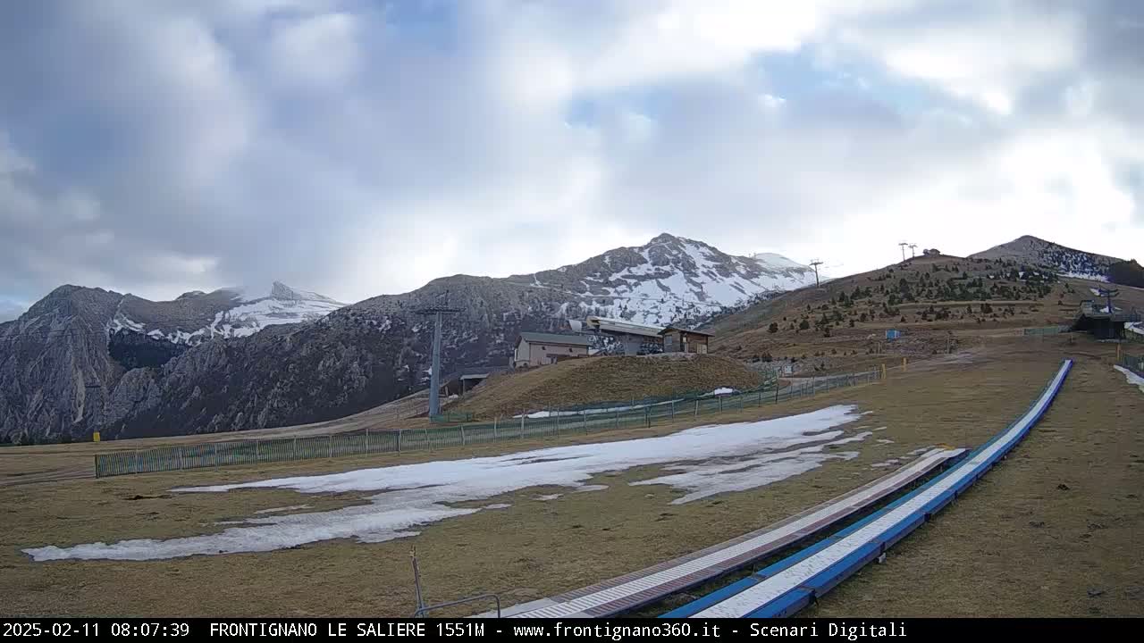 A partly cloudy day reveals snow-capped mountains, a ski lift, and a grassy area with patches of snow and two long, blue ramps.
