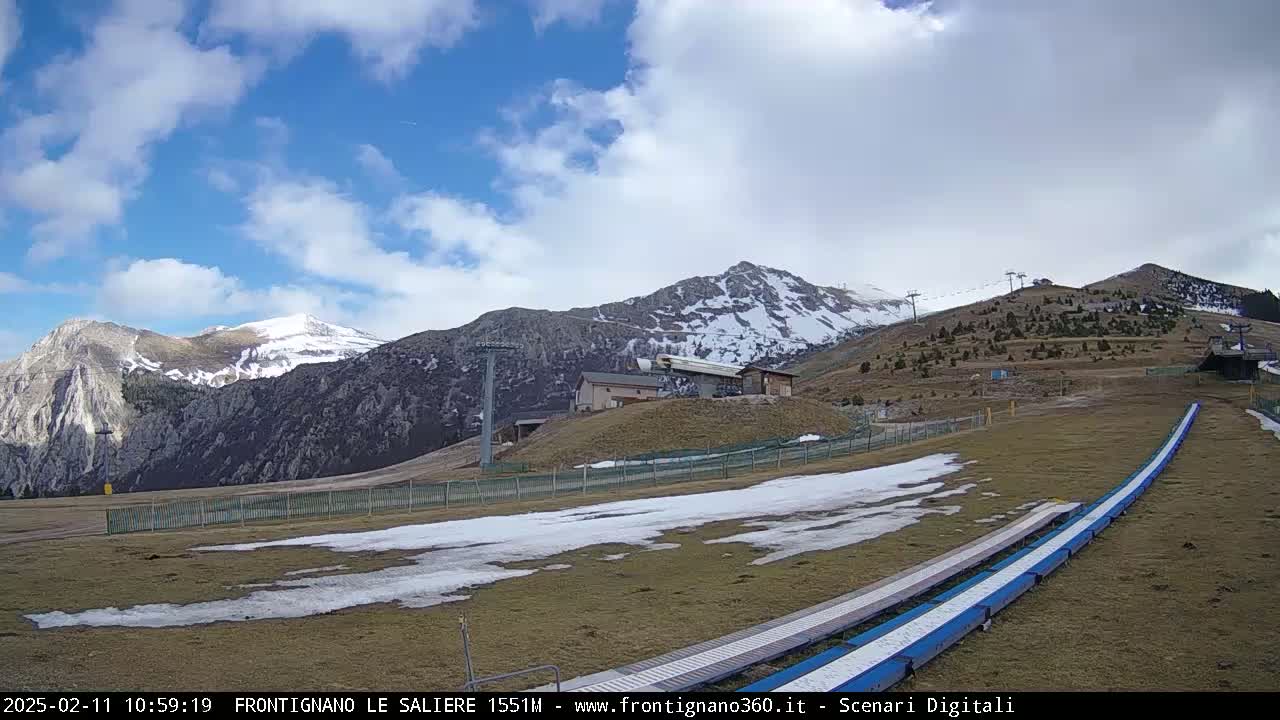 A partly cloudy day shows snow-capped mountains with a ski lift and a grassy area with patches of snow and what appears to be a ski lift access ramp.