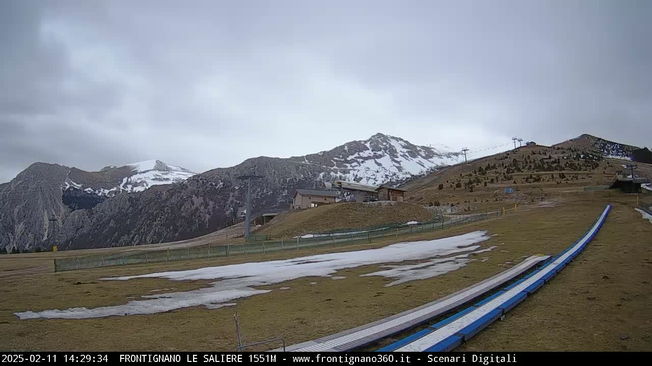 A mostly cloudy day reveals snow-dusted mountains and a ski resort with a few buildings and ski lifts, where patchy snow lies on the brown grass.