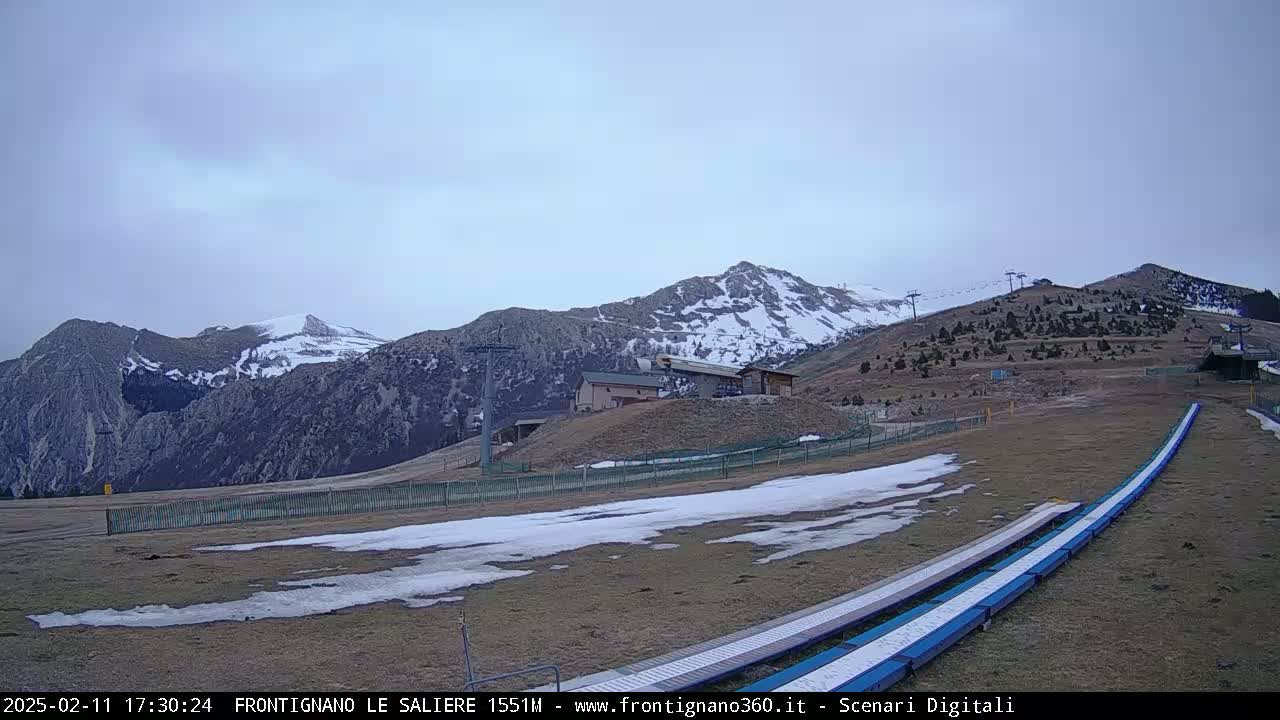 A ski slope with patches of snow sits below snow-capped mountains under an overcast sky.