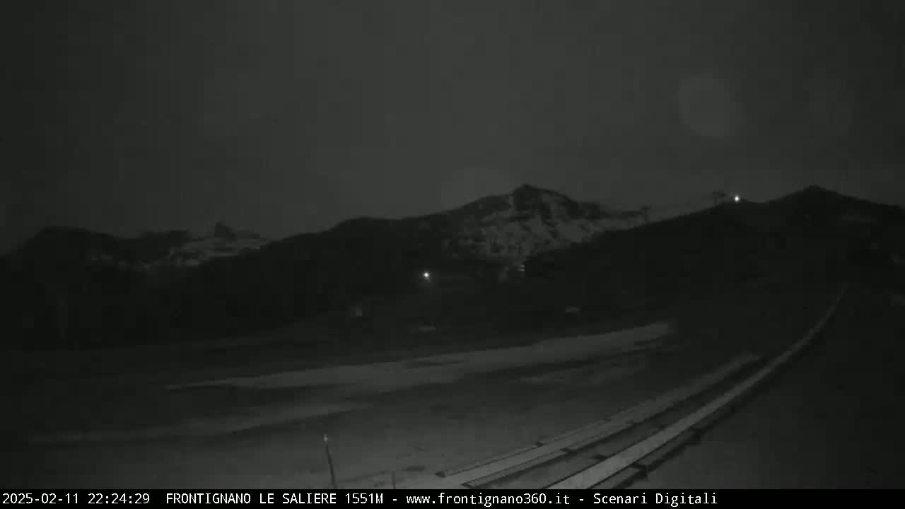 A nighttime, low-light view of a snow-dusted mountain range, with empty bleachers in the foreground.