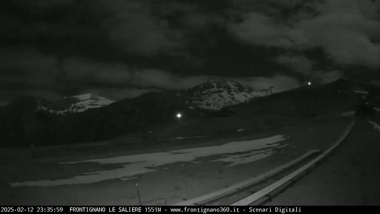 A nighttime, cloudy view of snow-covered mountains and a mostly snow-covered ground, with a road and some lights visible in the foreground.